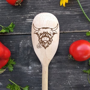 Puede incluir: Una cuchara de madera con un dise&ntilde;o de vaca Highland marr&oacute;n. La cuchara est&aacute; sobre una superficie de madera con pimientos rojos y hierbas verdes.