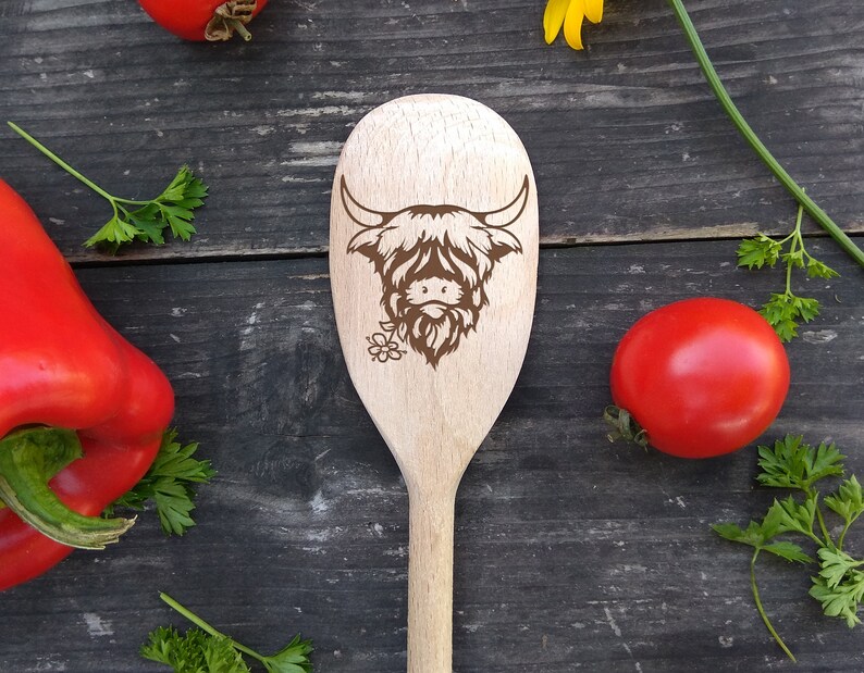 Puede incluir: Una cuchara de madera con un dise&ntilde;o de vaca Highland marr&oacute;n. La cuchara est&aacute; sobre una superficie de madera con pimientos rojos y hierbas verdes.
