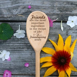May include: A wooden spoon with the text "Friends are the most important ingredient in the recipe of life! Friendsgiving 2026." The spoon is surrounded by flowers and leaves on a wooden surface.