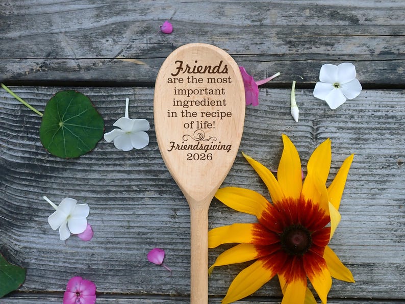 May include: A wooden spoon with the text "Friends are the most important ingredient in the recipe of life! Friendsgiving 2026." The spoon is surrounded by flowers and leaves on a wooden surface.