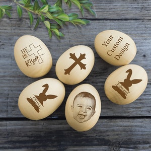 Könnte beinhalten: Sechs hölzerne Ostereier mit verschiedenen Designs, darunter ein Kreuz, ein Hasenkaninchen, ein Foto eines Babys und der Text "He is Risen".