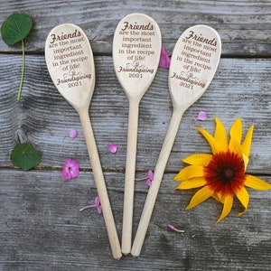 May include: Three wooden spoons with engraved text: "Friends are the most important ingredient in the recipe of life! Friendsgiving 2021." The spoons are arranged on a weathered wooden surface, with a yellow and red flower and scattered petals.