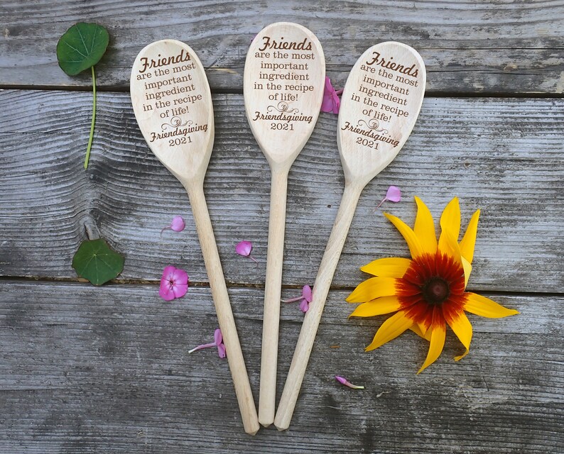 May include: Three wooden spoons with engraved text: "Friends are the most important ingredient in the recipe of life! Friendsgiving 2021." The spoons are arranged on a weathered wooden surface, with a yellow and red flower and scattered petals.