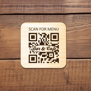 Puede incluir: Un posavasos cuadrado de madera clara con esquinas redondeadas que presenta un c&oacute;digo QR grabado con l&aacute;ser en color marr&oacute;n oscuro. El texto SCAN FOR MENU est&aacute; impreso sobre el c&oacute;digo y las palabras Bar & Cafe est&aacute;n centradas.