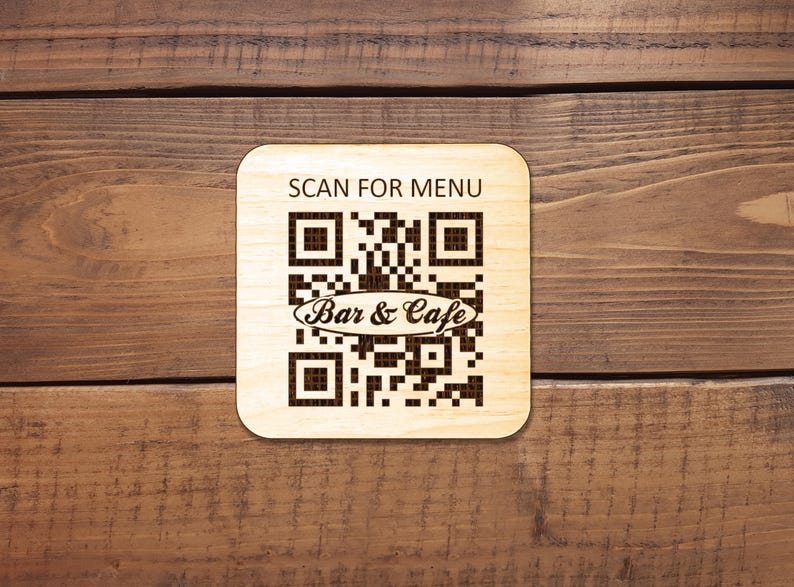 Puede incluir: Un posavasos cuadrado de madera clara con esquinas redondeadas que presenta un c&oacute;digo QR grabado con l&aacute;ser en color marr&oacute;n oscuro. El texto SCAN FOR MENU est&aacute; impreso sobre el c&oacute;digo y las palabras Bar & Cafe est&aacute;n centradas.