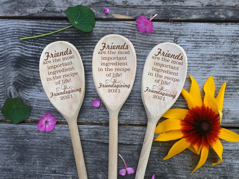 May include: Three wooden spoons with engraved text: "Friends are the most important ingredient in the recipe of life!" and "Friendsgiving 2021." The spoons are arranged on a rustic wooden surface, with flowers and leaves.