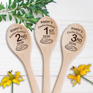 May include: Three light-colored wooden spoons featuring laser-engraved text and graphics. Each spoon displays 'Kipper's Cookie Contest', a place ranking (1st, 2nd, or 3rd), the year 2026, and a small illustration of a plate with three cookies.