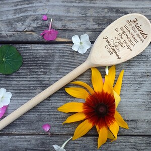 May include: A wooden spoon with the text "Friends are the most important ingredient in the recipe of life! Friendsgiving 2022". The spoon is surrounded by flowers and rests on a weathered wooden surface.