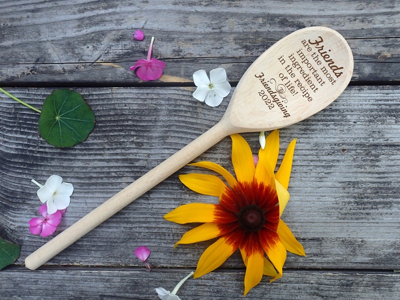May include: A wooden spoon with the text "Friends are the most important ingredient in the recipe of life! Friendsgiving 2022". The spoon is surrounded by flowers and rests on a weathered wooden surface.