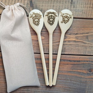 Puede incluir: Tres cucharas de madera con un dise&ntilde;o de vaca quemado en la parte delantera de cada cuchara. Las cucharas est&aacute;n dispuestas en fila y est&aacute;n sobre una superficie de madera. Una bolsa de tela blanca est&aacute; a la izquierda de las cucharas.
