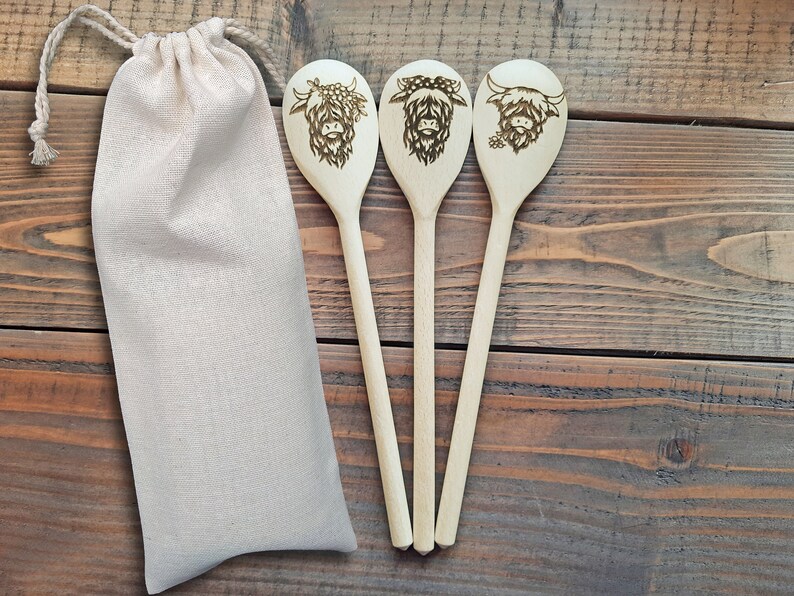 Puede incluir: Tres cucharas de madera con un dise&ntilde;o de vaca quemado en la parte delantera de cada cuchara. Las cucharas est&aacute;n dispuestas en fila y est&aacute;n sobre una superficie de madera. Una bolsa de tela blanca est&aacute; a la izquierda de las cucharas.
