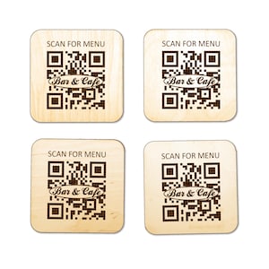 Puede incluir: Cuatro posavasos cuadrados de madera clara, cada uno con un c&oacute;digo QR grabado a l&aacute;ser en color marr&oacute;n oscuro. Sobre el c&oacute;digo QR, el texto dice SCAN FOR MENU, y centrado dentro del c&oacute;digo QR aparece el texto Bar & Cafe en cursiva.