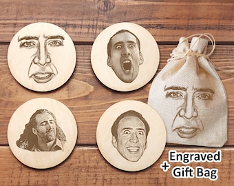 May include: Set of four wooden coasters featuring different images of Nicolas Cage's face. The coasters are round and have a natural wood finish. The set comes with a small burlap bag with an image of Nicolas Cage's face on it. The bag has the text "Engraved + Gift Bag" printed on it.