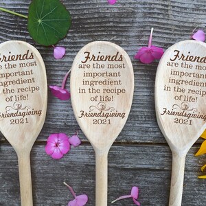 May include: Three wooden spoons with engraved text: "Friends are the most important ingredient in the recipe of life!" and "Friendsgiving 2021." The spoons are arranged on a wooden surface, decorated with flowers and leaves.