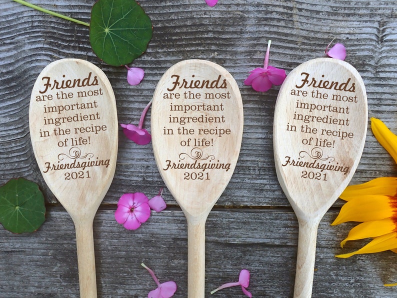 May include: Three wooden spoons with engraved text: "Friends are the most important ingredient in the recipe of life!" and "Friendsgiving 2021." The spoons are arranged on a wooden surface, decorated with flowers and leaves.