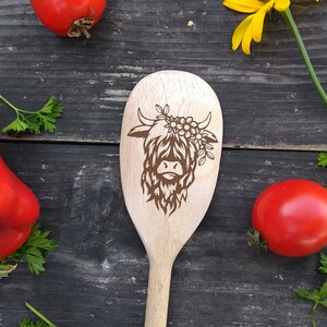 Puede incluir: Una cuchara de madera con un dise&ntilde;o de vaca de las tierras altas. La vaca es marr&oacute;n y lleva una corona de flores. La cuchara est&aacute; sobre un fondo de madera.