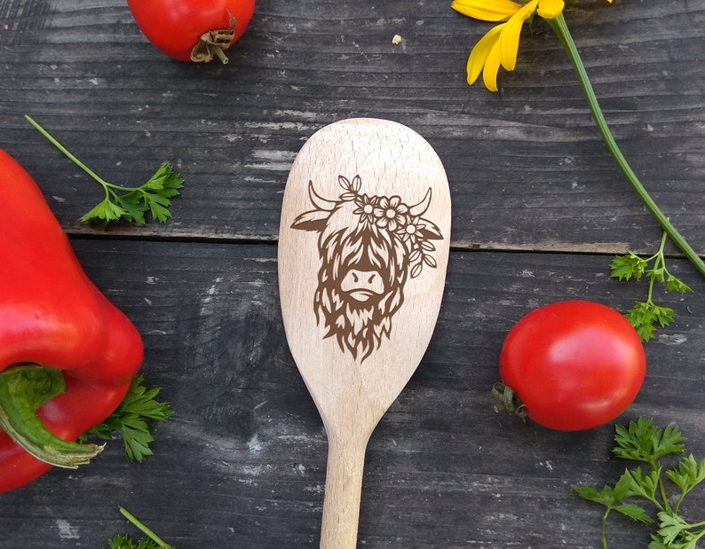 Puede incluir: Una cuchara de madera con un dise&ntilde;o de vaca de las tierras altas. La vaca es marr&oacute;n y lleva una corona de flores. La cuchara est&aacute; sobre un fondo de madera.