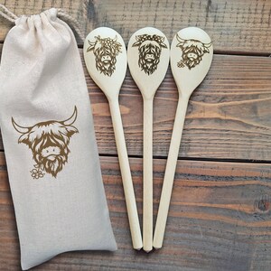 Puede incluir: Tres cucharas de madera con un dise&ntilde;o de vaca de las tierras altas, cada una con una expresi&oacute;n diferente. Las cucharas est&aacute;n en una bolsa de lona blanca.