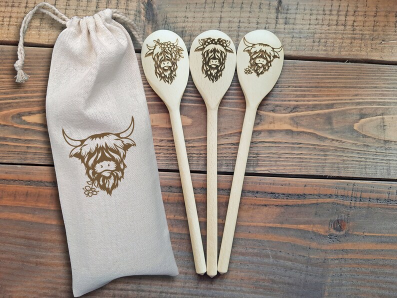 Puede incluir: Tres cucharas de madera con un dise&ntilde;o de vaca de las tierras altas, cada una con una expresi&oacute;n diferente. Las cucharas est&aacute;n en una bolsa de lona blanca.