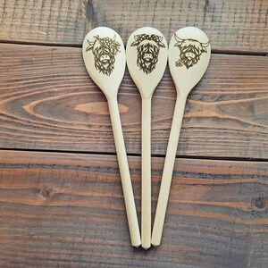Puede incluir: Tres cucharas de madera con un dise&ntilde;o de vaca Highland quemado en la parte delantera de cada cuchara. Las cucharas est&aacute;n dispuestas en fila sobre una superficie de madera.