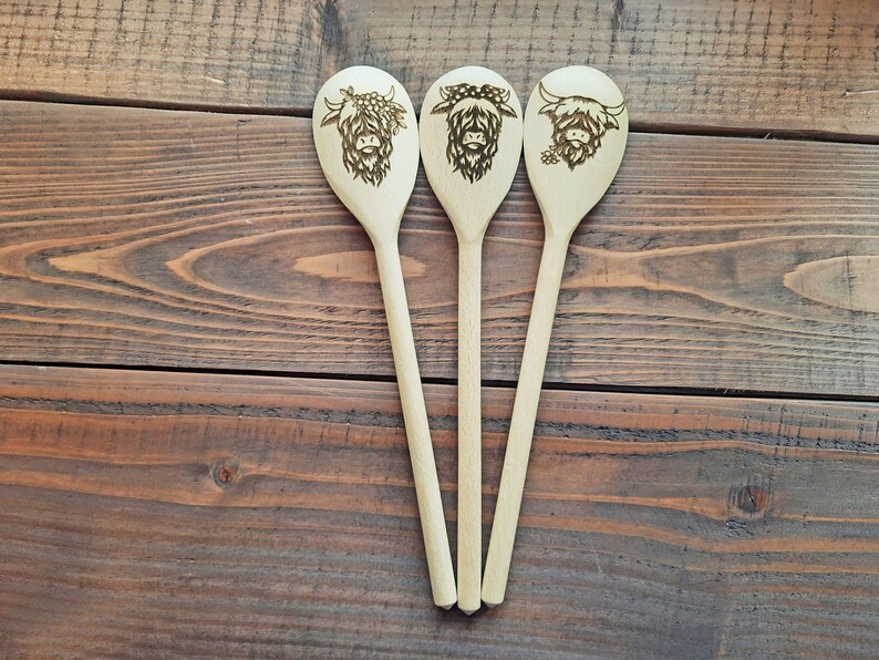 Puede incluir: Tres cucharas de madera con un dise&ntilde;o de vaca Highland quemado en la parte delantera de cada cuchara. Las cucharas est&aacute;n dispuestas en fila sobre una superficie de madera.