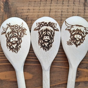 Puede incluir: Tres cucharas de madera con un dise&ntilde;o de vaca Highland quemado en la parte delantera de cada cuchara. La vaca de la cuchara de la izquierda lleva una corona de flores, la cuchara del medio tiene un lazo de lunares y la vaca de la cuchara de la derecha tiene una flor detr&aacute;s de la oreja.