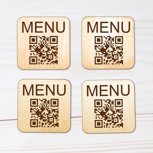 Puede incluir: Cuatro posavasos cuadrados de madera clara con esquinas redondeadas. Cada posavasos presenta la palabra MENU impresa en letras may&uacute;sculas de color marr&oacute;n oscuro sobre un c&oacute;digo QR marr&oacute;n oscuro. La veta de la madera es visible en cada pieza.