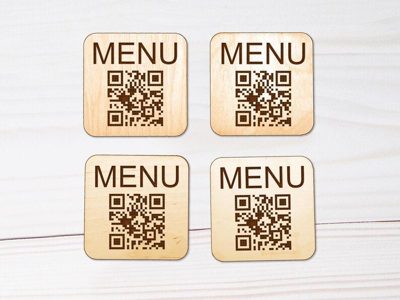 Puede incluir: Cuatro posavasos cuadrados de madera clara con esquinas redondeadas. Cada posavasos presenta la palabra MENU impresa en letras may&uacute;sculas de color marr&oacute;n oscuro sobre un c&oacute;digo QR marr&oacute;n oscuro. La veta de la madera es visible en cada pieza.