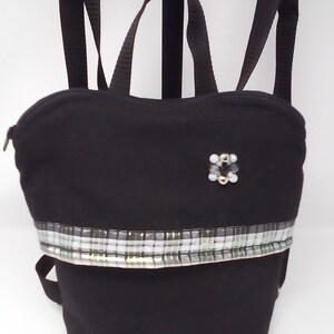 Peut inclure: Un sac à dos en toile noire avec une bordure en tissu à carreaux argent et blanc et un petit ornement en perles. Le sac à dos a des bretelles réglables.