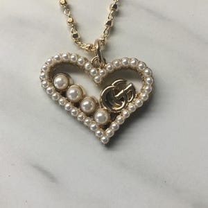 May include: Gold-toned heart-shaped pendant necklace with a pearl-lined border and a gold double-G logo. The pendant features five pearls and hangs from a delicate gold chain. A stylish accessory.