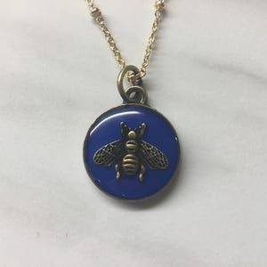 May include: A gold-colored necklace with a round pendant featuring a blue background and a detailed bronze-colored bee. The necklace has a delicate chain and a simple design, suitable for everyday wear.