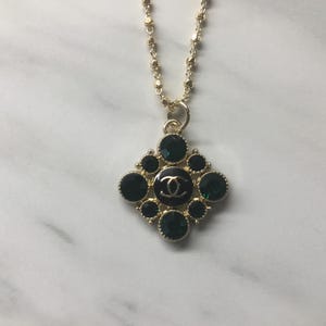 May include: Gold-toned necklace with a pendant featuring a black and gold logo surrounded by dark green gemstones. The pendant is a diamond shape. The necklace has a delicate chain.