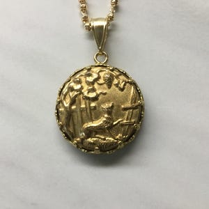 May include: A gold-colored pendant necklace with a detailed circular pendant. The pendant features a relief scene of a deer in a forest setting. The necklace chain is also gold-colored.