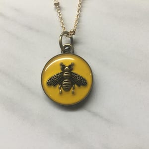 May include: A gold-colored necklace with a round pendant featuring a yellow background and a detailed bee design. The bee is dark brown with outstretched wings. The necklace is on a white and gray marble surface.
