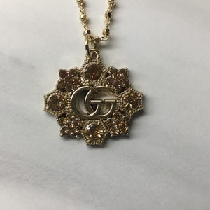 May include: Gold-toned necklace with a pendant featuring a floral design and the interlocking "GG" logo. The pendant is adorned with brown-toned crystals. The necklace is on a gold chain.