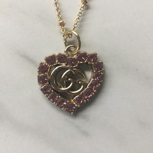 May include: Gold-toned necklace featuring a heart-shaped pendant. The pendant is adorned with pink gemstones and a double-G logo in the center. The necklace has a delicate chain.