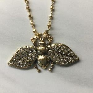 May include: Gold-toned bee pendant necklace. The bee pendant features a detailed design with crystal accents on the wings and body. The necklace has a delicate chain with small, round beads.