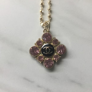 May include: Gold-tone necklace with a pendant featuring a black center with a double C logo, surrounded by pink gemstones. The pendant is diamond-shaped. The necklace is on a white marble surface.