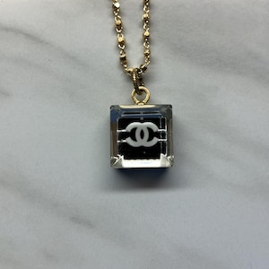 May include: A gold-toned chain necklace with a square pendant. The pendant is clear with a black background and a white double-C logo. The necklace is displayed on a marble surface.