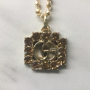 May include: Gold-toned necklace featuring a square pendant with a double-G logo at the center, surrounded by brown gemstones. The chain is a delicate beaded design, and the pendant hangs against a white background.