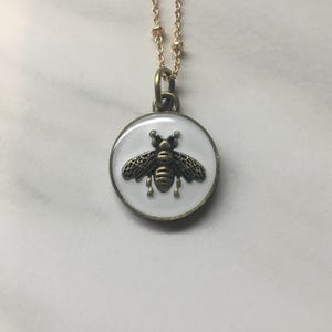 May include: A gold-toned necklace featuring a round pendant with a white background. The pendant showcases a detailed, antique-style bee design in shades of bronze and black. The necklace chain is delicate and gold.