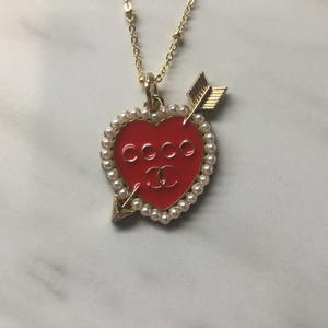 May include: Gold-toned necklace with a red heart pendant. The heart is pierced by a gold arrow and encircled by faux pearls. The word "COCO" and a double "C" logo are visible on the heart.
