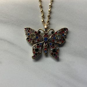 May include: Gold-toned butterfly pendant necklace. The butterfly pendant is encrusted with colorful rhinestones in red, blue, black, and clear. The necklace chain is a delicate gold color.
