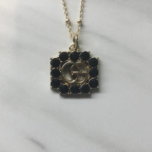 May include: Gold-toned necklace with a square pendant featuring a gold double-G logo surrounded by black gemstones. The chain is delicate and the pendant is centered in the frame, showcasing the jewelry's design.