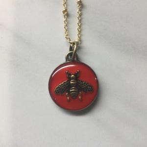 May include: A gold-colored chain necklace with a round pendant. The pendant features a red background with a detailed bronze-colored bee design. The necklace is a delicate piece of jewelry.
