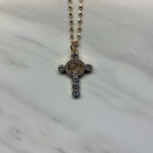 May include: A gold-colored necklace with a cross pendant. The cross is adorned with clear, sparkling crystals and a circular design at the center. The chain is delicate and has a subtle, textured pattern.