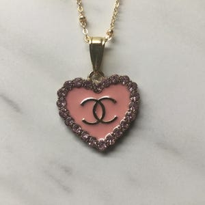 May include: A gold-toned necklace featuring a heart-shaped pendant. The pendant has a pink enamel base with a double-C logo in the center, surrounded by small, sparkling pink crystals. The necklace hangs on a delicate gold chain.