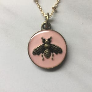 May include: A gold-toned necklace featuring a round pendant with a pink enamel background. The pendant showcases a detailed, antique-style bee design in a darker metallic finish. The necklace has a delicate chain.
