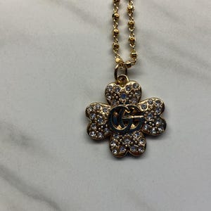 May include: A gold-colored necklace with a four-leaf clover pendant. The pendant is encrusted with small, clear crystals and features a double-G logo in the center. The chain is a delicate beaded style.