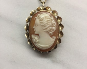 May include: A vintage cameo pendant necklace. The oval pendant features a carved profile of a woman in white against a peach background, set in a gold-tone frame with a scalloped edge. The necklace has a gold-tone chain.